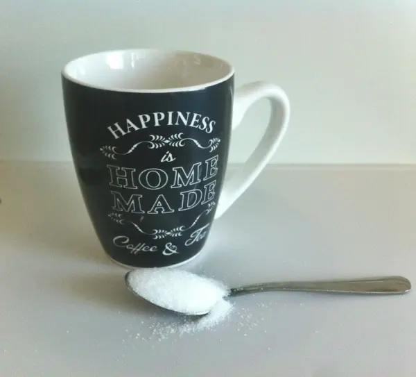 Eine schwarze Tasse steht auf einem Tisch, während ein Löffel mit Zucker daneben liegt, bereit zum Umrühren.