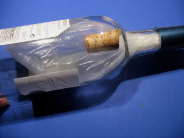 Eine Flasche steht auf einem blauen Untergrund, der Korken wird mit einem Kunststoffüberzug entfernt.