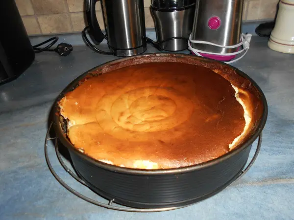 Ein Käsekuchen mit goldenem, leicht angebranntem Rand steht in einer Backform auf einer Küchenplatte, umgeben von Gerätschaften.