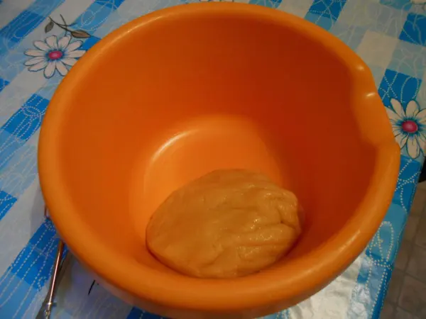 Ein orangefarbener Schüssel enthält einen Teigball, der zur Zubereitung eines Käsekuchens vorbereitet wird, auf einem gemusterten Tisch.