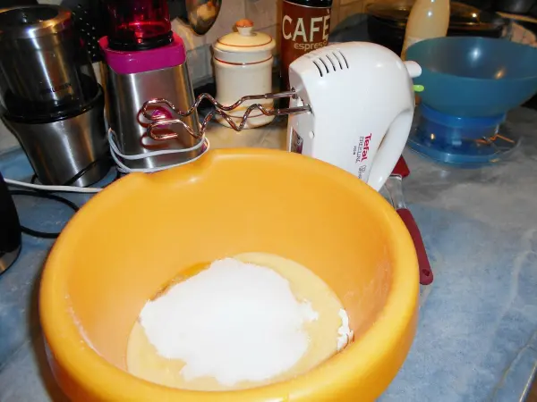 In einer orangefarbenen Schüssel wird Zucker mit einem Handmixer verrührt, um einen einfachen Käsekuchen zuzubereiten.