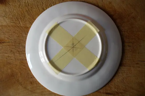 Ein Teller aus altem Porzellan liegt auf einer Holzoberfläche, mit einem X aus Klebeband auf der Rückseite für die Etagere-Befestigung.