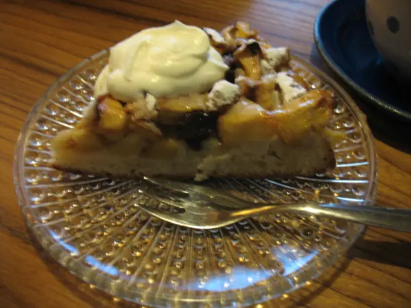 Ein Stück Apfeltarte liegt auf einem gläsernen Dessertteller, garniert mit Sahne, auf einem Holztisch.