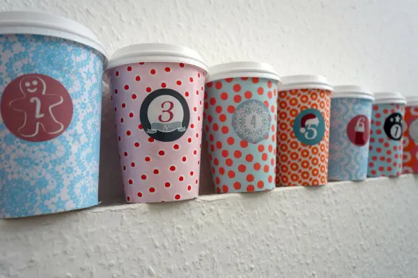 Bunte Coffee-to-go-Becher stehen an einer Wand, dekoriert mit Zahlen für einen Adventskalender.