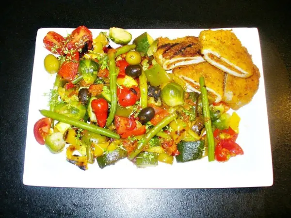 Pesto-Putenschnitzel liegt neben buntem, knackigem Ofengemüse auf einem weißen Teller, umgeben von einem dunklen Hintergrund.
