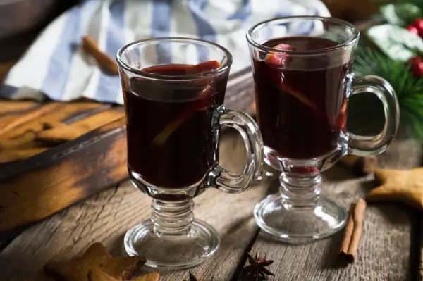 Zwei Gläser mit heißem Glühwein stehen auf einem rustikalen Holztisch, umgeben von Zimtstangen und Weihnachtsdekoration.