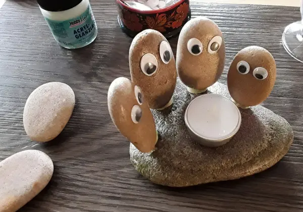 Fünf bemalte Steine mit Augen um einen kleinen Kerzenhalter auf einem Tisch versammeln sich; fröhliche Atmosphäre.