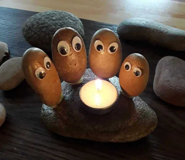 Fünf mit Wackelaugen versehene Steine sitzen um eine brennende Kerze auf einem Tisch, teilen einen gemütlichen Moment.