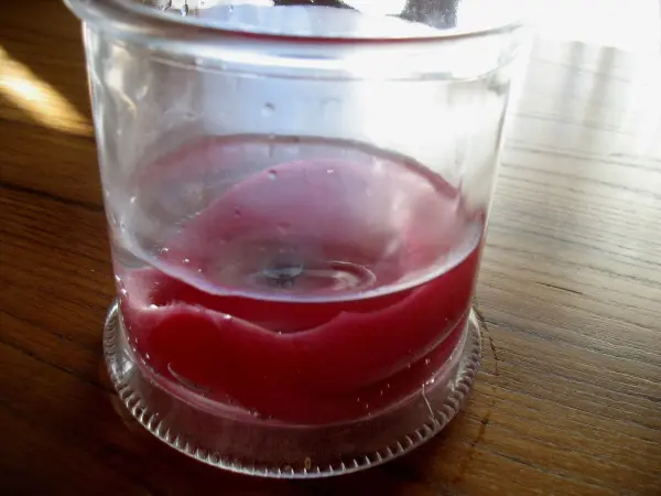 Ein transparentes Glasgefäß zeigt geschmolzenes Wachs in roter Farbe, das im Sonnenlicht auf einem Holztisch ruht.