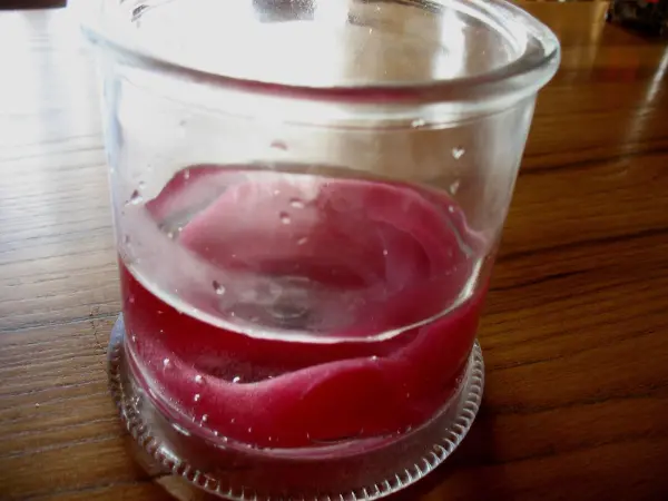 Ein durchscheinendes Glasgefäß steht auf einem Holztisch, gefüllt mit geschmolzenem Wachs in rötlicher Farbe.