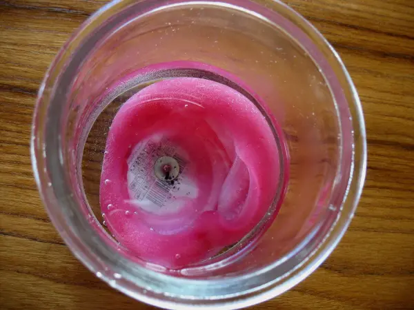 Im Glasgefäß schwimmt Wachs, das sich dabei auflöst und entfaltet eine rosa Farbe, auf einem Holzuntergrund.