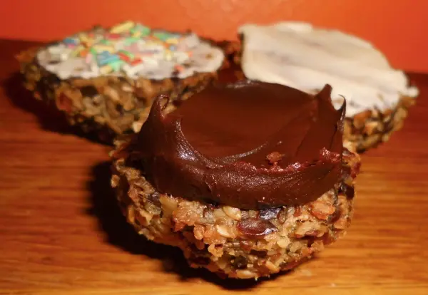 Haferflocken-Leinsamen-Brötchen liegen appetitlich auf einem Holzbrett, zwei sind mit Zuckerguss dekoriert, einer mit Schokoladencreme.
