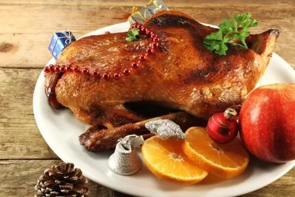 Auf einem weißen Teller liegt eine Weihnachtsente, dekoriert mit einer roten Perlenkette und umgeben von Orangen und Äpfeln.
