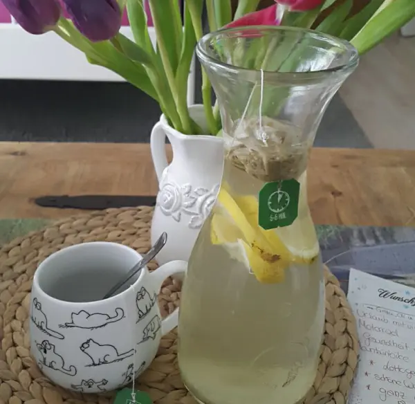 Ein Glas mit grünem Tee, frischen Zitronenscheiben und Ingwer steht auf einem Tisch, umgeben von bunten Tulpen.