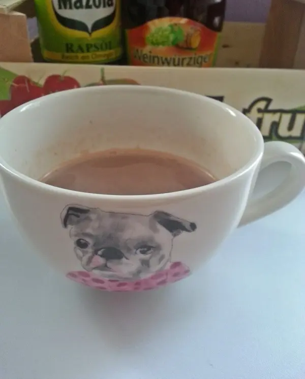 Eine Tasse heiße Schokolade steht auf einem Tisch, dekoriert mit einem Hundemotiv und umgeben von Getränkeflaschen.