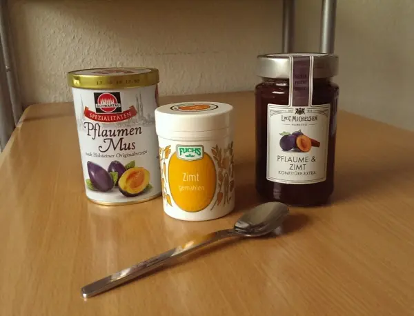 Drei Zutaten stehen auf einem Holzregal: Pflaumenmus, Zimt und ein Glas Marmelade. Ein Löffel liegt bereit zum Mischen.