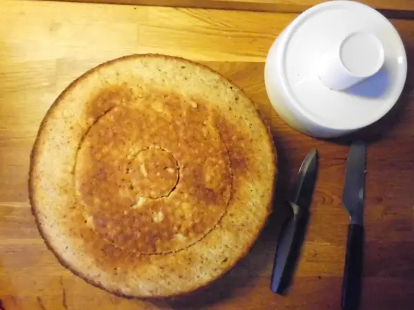 Eine runde Nusstorte liegt auf einem Holztisch, daneben stehen ein Schüssel und Besteck bereit zur Dekoration.