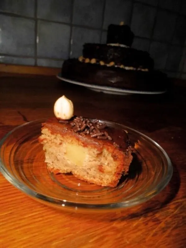 Ein Stück Nusstorte liegt auf einem Glasuntersetzer, während im Hintergrund eine dreistöckige Torte sichtbar ist.