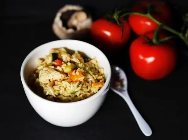 Ein gefüllter Schüssel mit Reisgericht, Pute und buntem Gemüse steht vor roten Tomaten auf einem dunklen Hintergrund.