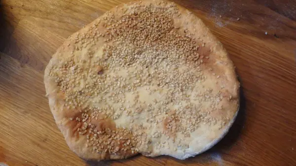Ein rundes Pfannenbrot mit einer goldbraunen, leicht knusprigen Oberfläche liegt auf einem Holzbrett und ist mit Sesampaste bestreut.