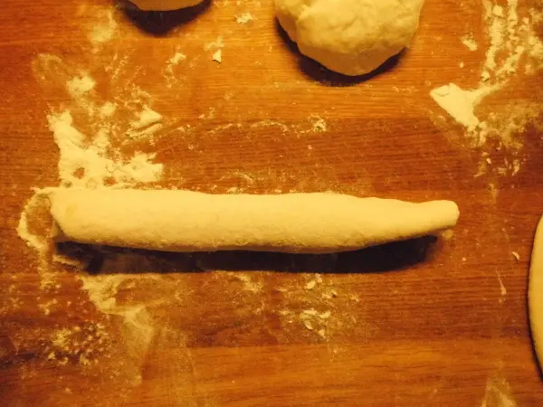 Ein länglicher Teigstrang liegt auf einer bemehlten Holzfläche, umgeben von weiteren Teigkugeln für das Pfannenbrot.