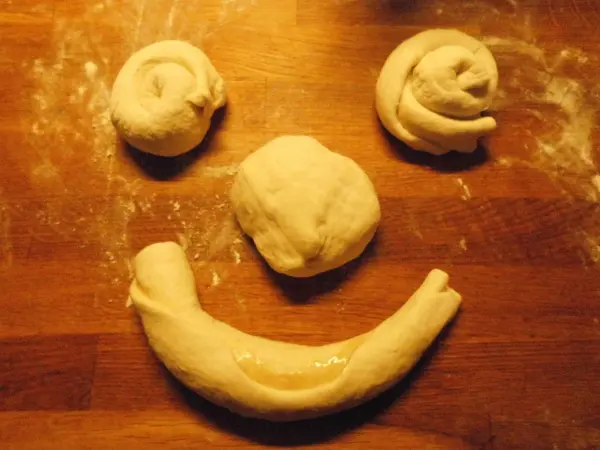 Auf einer Holzplatte liegen Teigstücke in verschiedenen Formen, die zusammen ein lächelndes Gesicht bilden.