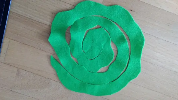 Ein grüner, spiralförmiger Osterschmuck liegt auf einem Holzboden und könnte als Dekoration für einen österlichen Kranz dienen.