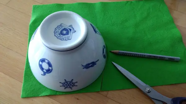 Ein weißer Teller mit blauen Mustern liegt kopfüber auf einem grünen Tuch, daneben eine Schere und ein Stift.