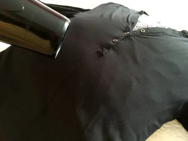 Ein schwarzes Hemd liegt flach, während ein Haartrockner direkt darauf gerichtet ist, um es ohne Bügeleisen zu glätten.