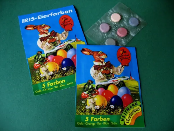 Bunte Ostereier-Färbesets liegen auf einem grünen Tisch, umgeben von Farbbäcken und einer fröhlichen Hasenillustration.