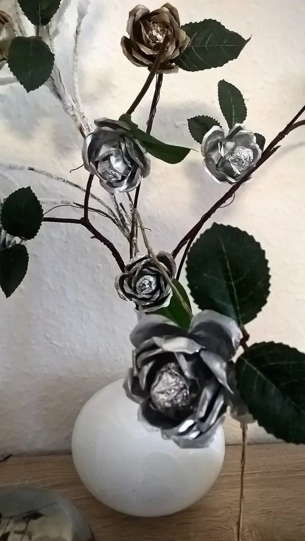 Silberne Rosen aus Teelichtschalen stehen in einer weißen Vase, umgeben von grünen Blättern und Zweigen.