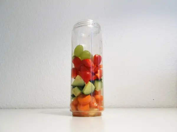 Ein transparenter Smoothie Mixer ist gefüllt mit bunten Früchten wie Melonen, Trauben und Gurken, vor einem neutralen Hintergrund.