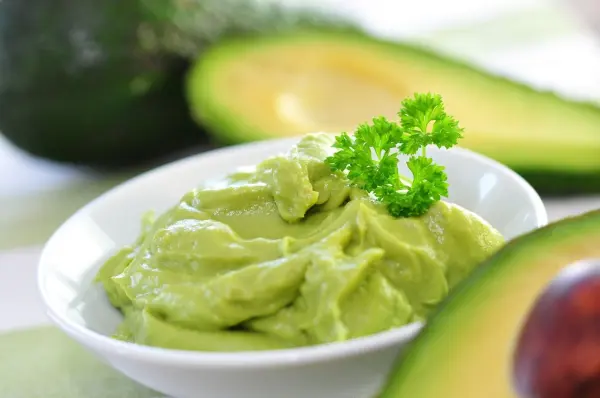 Ein cremiger Avocado-Dip liegt in einer weißen Schüssel, umgeben von frischen Avocados auf einem hellen Tisch.