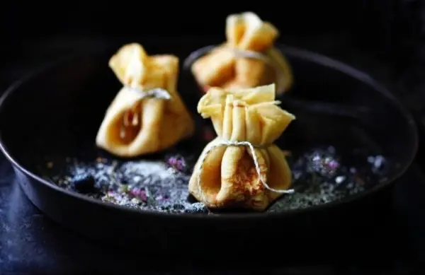 Drei gefüllte Pfannkuchen-Zitronen-Säckchen liegen auf einer schwarzen Pfanne, umgeben von Puderzucker.