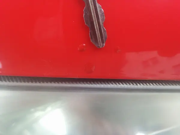 Eine rote Autokarosserie zeigt einen kleinen Steinschlag in der Lackierung mit metallischem Hintergrund.