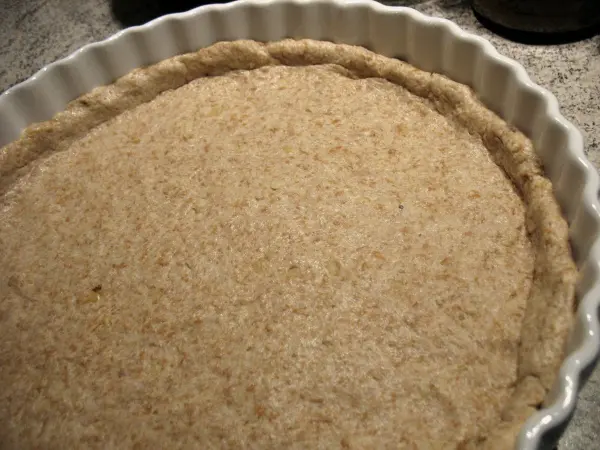 Eine ungebackene Tarteboden aus Rührteig liegt in einer weißen Tarteform auf einer Arbeitsfläche, bereit für Käse und Spargel.