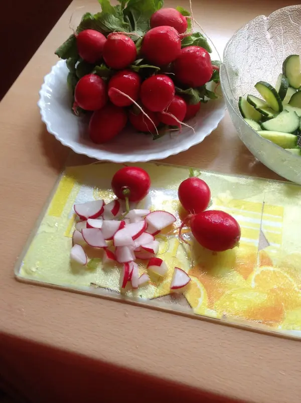 Frische Radieschen liegen auf einem Tisch, während einige bereits in Scheiben geschnitten sind. Neben ihnen ist eine Schüssel mit Gurken.