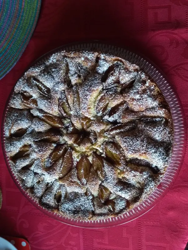 Ein runder Pflaumenkuchen liegt auf einem roten Tisch, mit Puderzucker bestäubt und saftigen Pflaumen in kunstvollem Muster.