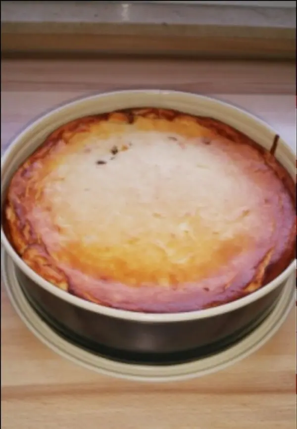 Ein käsekuchen mit goldbrauner Oberfläche steht in einer runden Form auf einem Holzuntergrund, frisch gebacken und appetitlich.