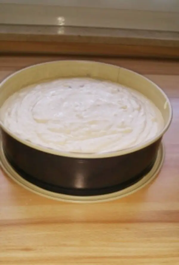 Ein Käsekuchen ohne Boden steht in einer runden Form auf einer Holzoberfläche und wartet darauf, gebacken zu werden. Saftiger Apfelmus-Fleck ist sichtbar.