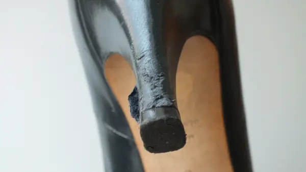 Ein schwarzer High Heel steht im Fokus, der an der Spitze beschädigt ist. Die Umgebung ist neutral und hell.
