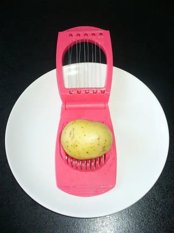 Eine Kartoffel liegt in einem rosa Schnneider auf einem weißen Teller. Der Schneider schneidet die Pellkartoffeln in Scheiben.