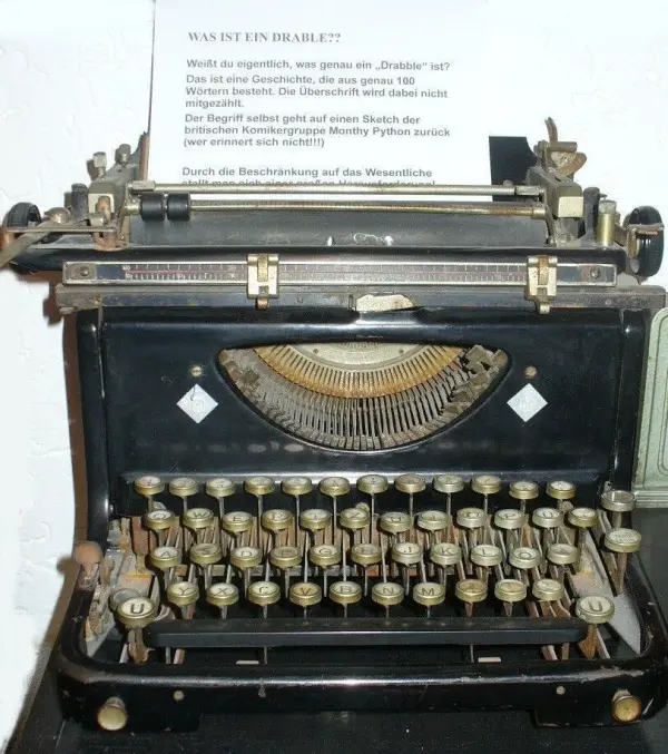 Eine alte Schreibmaschine steht auf einem Tisch. Sie wird nicht benutzt, während ein Text über das Schreiben von Drabbles im Hintergrund sichtbar ist.