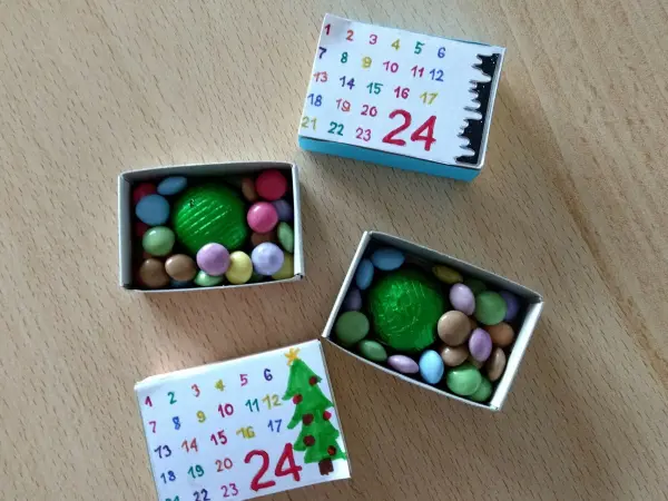 Mini-Adventskalender aus Streichholzschachteln mit bunten Süßigkeiten und einem grünen Ball im Inneren, auf einem Tisch.