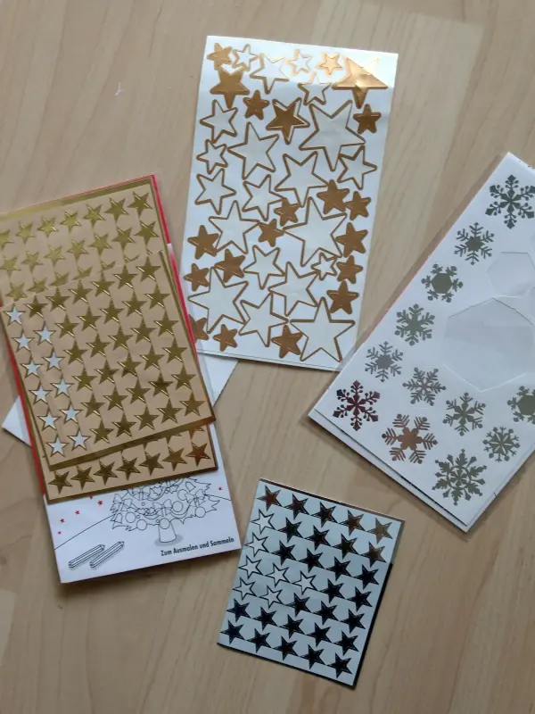 Verschiedene Aufkleber mit goldenen und silbernen Sternen sowie Schneeflocken liegen auf einem hellen Tisch, ideal für einen Mini-Adventskalender.