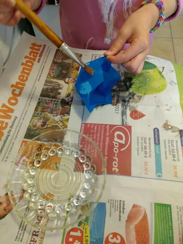 Eine Person bemalt einen blauen Papier-Windlichter auf einer Zeitung, während eine Glasplatte daneben liegt.