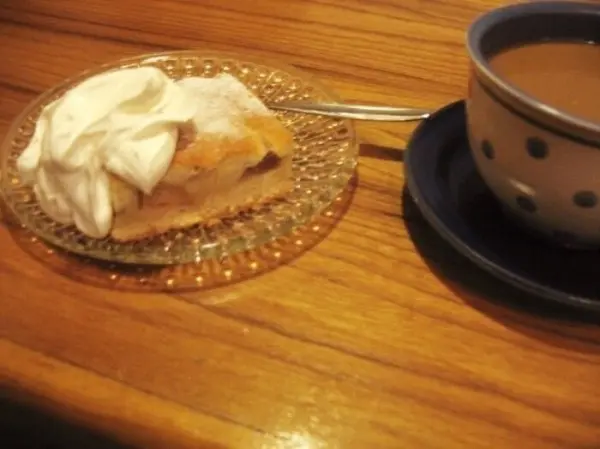 Ein Blechkuchen mit Sahne liegt auf einem gläsernen Teller neben einer Tasse Kaffee auf einem Holztisch.