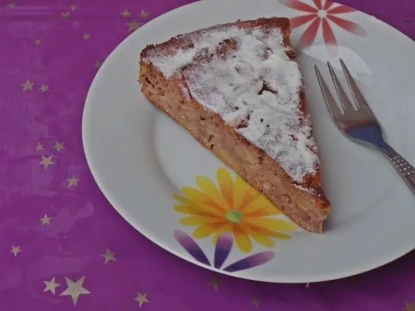 Ein Stück Apfelkuchen mit Kastanienmehl und Eiweißpulver liegt auf einem blumengemusterten Teller auf einem lila Tisch.