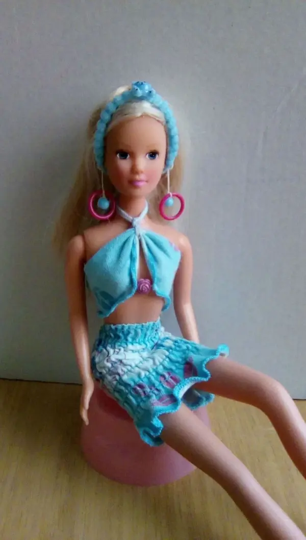 Eine Barbie-Puppe trägt blaue Kleidung und große Ohrringe, während sie auf einem rosa Hocker sitzt.