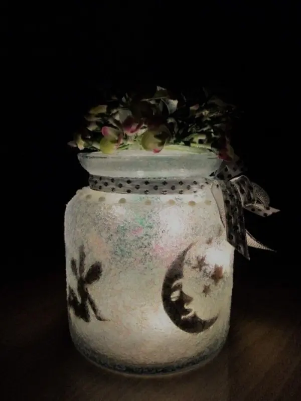 Ein transparentes Windlicht mit bemalten Motiven von Mond und Sternen leuchtet sanft im Dunkeln, umgeben von einer blühenden Deko.